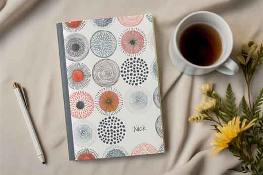 a notebook with a colorful, abstract design, a cup of coffee, a pen, and some flowers on a beige surface.